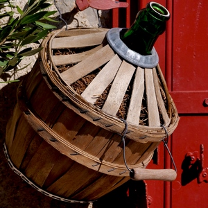 Dame Jeanne recouverte de bois accrochée à un mur en pierres devant une porte en bois rouge - France  - collection de photos clin d'oeil, catégorie clindoeil